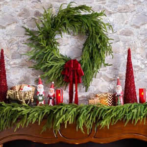 May include: A wooden mantel decorated for Christmas with a green garland, red velvet bows, and small Santa figurines. There are also two red sparkly cone-shaped Christmas trees and red candles.