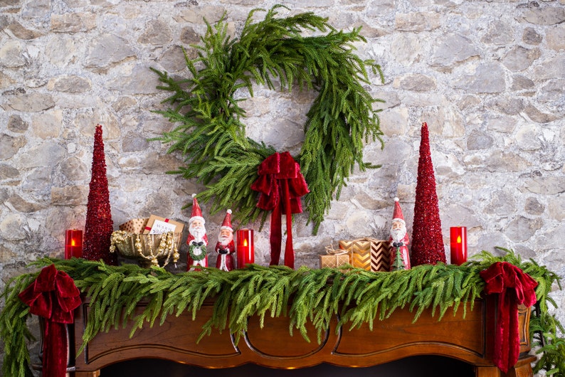 May include: A wooden mantel decorated for Christmas with a green garland, red velvet bows, and small Santa figurines. There are also two red sparkly cone-shaped Christmas trees and red candles.