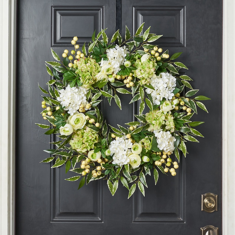 Peut inclure: Une couronne florale verte et blanche avec des hortensias blancs et des feuilles vertes. La couronne est d&eacute;cor&eacute;e de petites baies jaunes.