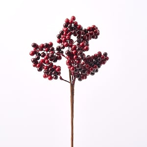 May include: A decorative floral pick featuring clusters of small, round berries in shades of deep crimson, bright red, and dark burgundy. The berries are attached to a central brown stem wrapped in a textured, paper-like material for a natural look.
