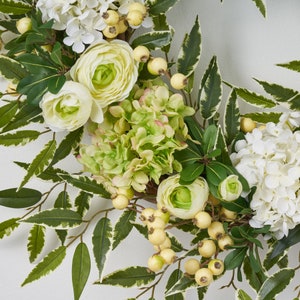 Peut inclure: Une couronne florale verte et blanche avec des hortensias blancs, des hortensias verts et des renoncules blanches. La couronne est d&eacute;cor&eacute;e de petites baies blanches.
