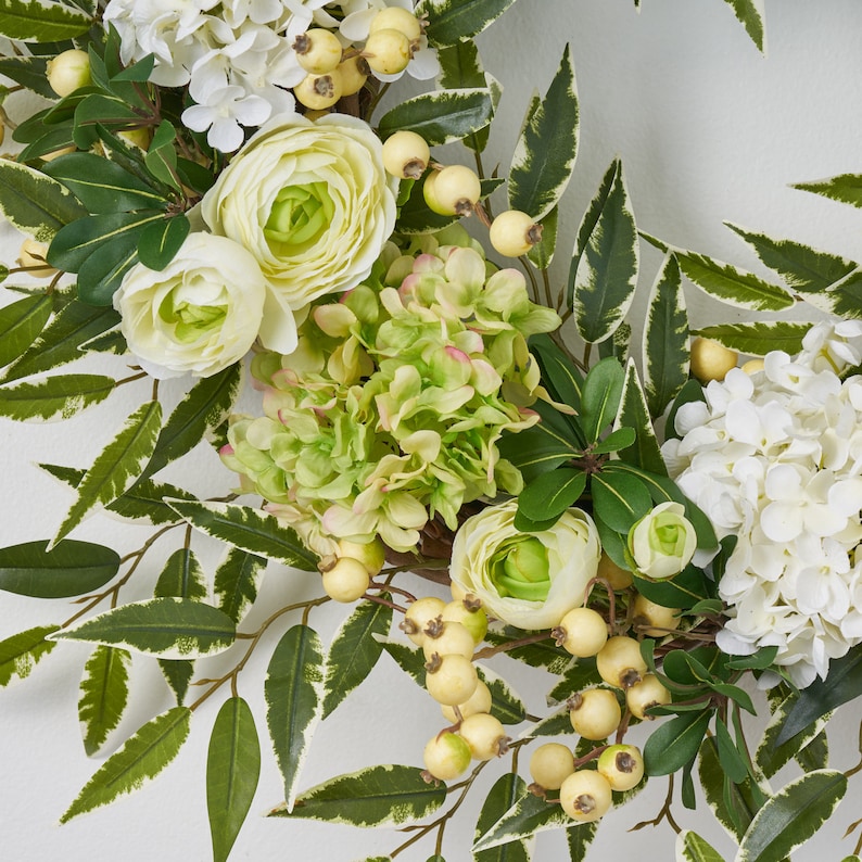 Peut inclure: Une couronne florale verte et blanche avec des hortensias blancs, des hortensias verts et des renoncules blanches. La couronne est d&eacute;cor&eacute;e de petites baies blanches.