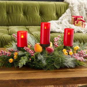 May include: A Christmas centerpiece with three red LED candles, each with a flickering flame. The candles are surrounded by greenery, berries, and fruit. The arrangement is on a wooden table. The candles are approximately 15 cm tall.