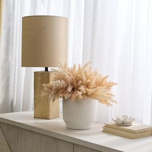 May include: A beige lamp with a linen shade sits on a stone base next to a white ceramic planter with dried pampas grass. The planter is on a light wood surface.
