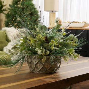 Puede incluir: Un centro de mesa decorativo con un arreglo de follaje artificial verde y plateado, que incluye agujas de pino, eucalipto y una piña. El arreglo está en una maceta gris-marrón texturizada con patrón de diamantes. Sobre una mesa de madera.