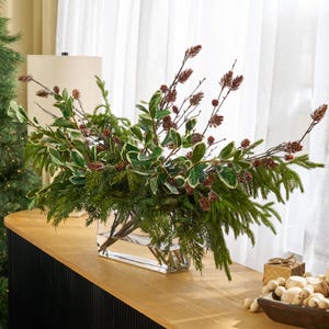 Op de afbeelding: Een rechthoekige glazen vaas gevuld met een arrangement van groen, waaronder dennennaalden, bonte bladeren en takken met kleine bruine dennenappels. De opstelling staat op een houten oppervlak.