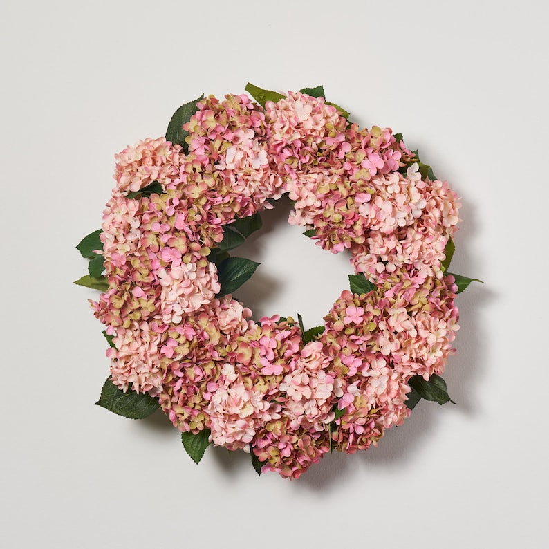 May include: A pink and green artificial hydrangea wreath. The wreath is made of pink hydrangea flowers and green leaves. The wreath is a circular shape and is approximately 12 inches in diameter.