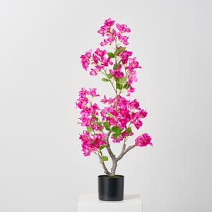 May include: A pink artificial bougainvillea tree in a black pot. The tree has a brown trunk and green leaves. The tree is on a white pedestal.