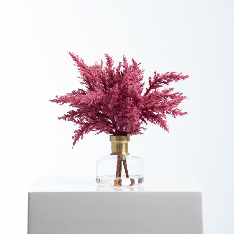 以下が含まれることがあります： 金色の蓋が付いた透明なガラスの花瓶に、ピンクのドライフラワーのブーケが活けられています。花瓶は白い表面の上に置かれています。