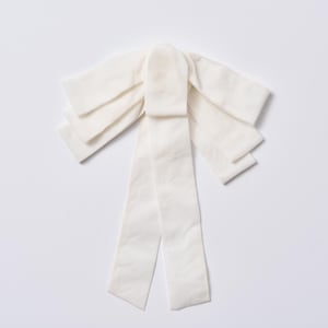 Puede incluir: Un lazo grande de color blanco roto hecho de tela suave. El lazo presenta múltiples bucles en capas y largas colas fluidas. El diseño es simétrico, con un nudo central y bucles radiantes. La tela tiene una textura sutil y la estética general es elegante y delicada.