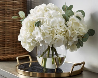 Arreglo floral grande de hortensias blancas y eucalipto en un jarrón de cristal redondo.