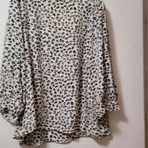 May include: A long-sleeved blouse with a leopard print pattern. The blouse is white with black and brown spots. The sleeves have a ruffled detail at the cuffs. The blouse is hanging on a clear plastic hanger.
