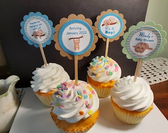 May include: Four cupcakes decorated with white frosting and colorful sprinkles. Each cupcake has a round, scalloped paper topper with a sloth illustration and text. The toppers read "Thanks for hanging out with us!", "Arriving... January 2022", "It's A Boy!" and "Mindy's Baby Shower".
