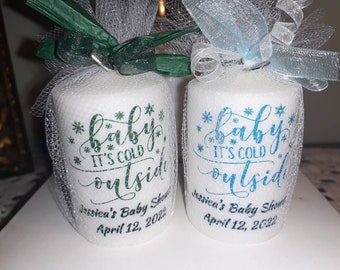May include: Two white cylindrical baby shower favors wrapped in netting and topped with bows. The favors are printed with the text "baby it's cold outside" and the date "April 12, 2022". One favor has green text and the other has blue text.
