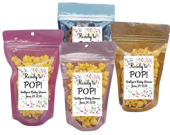 May include: Several clear plastic bags filled with popcorn. Each bag has a label that reads "Ready to POP!" and "Kaitlyn's Baby Shower June 24, 2026" with colorful confetti. The bags are in shades of pink, blue, and gold.
