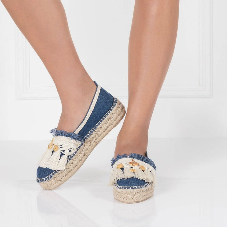 May include: A pair of blue denim espadrille shoes with white fringe and gold accents. The shoes have a flat sole and a comfortable fit.