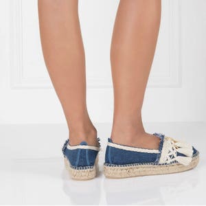 May include: A pair of blue denim espadrille shoes with white trim and tassels. The shoes have a jute sole and are worn by a person standing on a white surface.