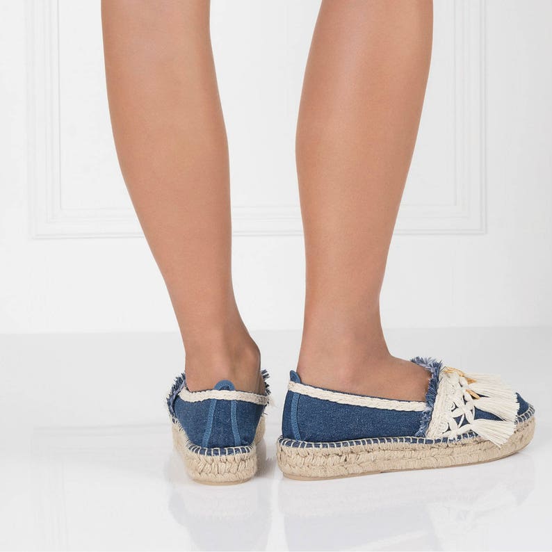 May include: A pair of blue denim espadrille shoes with white trim and tassels. The shoes have a jute sole and are worn by a person standing on a white surface.