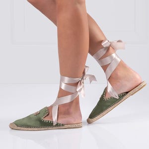 May include: Olive green espadrille flats with a jute sole and a satin ribbon tie. The flats have a rounded toe and a decorative stitched edge. The ribbon wraps around the ankle and ties in a bow.