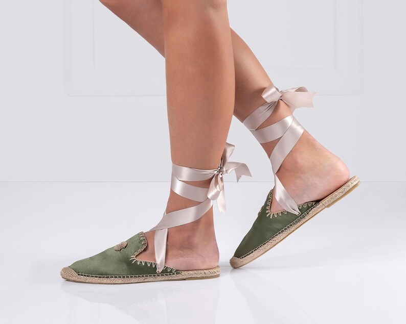 May include: Olive green espadrille flats with a jute sole and a satin ribbon tie. The flats have a rounded toe and a decorative stitched edge. The ribbon wraps around the ankle and ties in a bow.