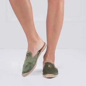 May include: Olive green satin slip-on mules with a woven jute sole. The shoes have a pointed toe and a small decorative emblem on the vamp. The edges are stitched with a contrasting thread. A stylish and comfortable footwear option.
