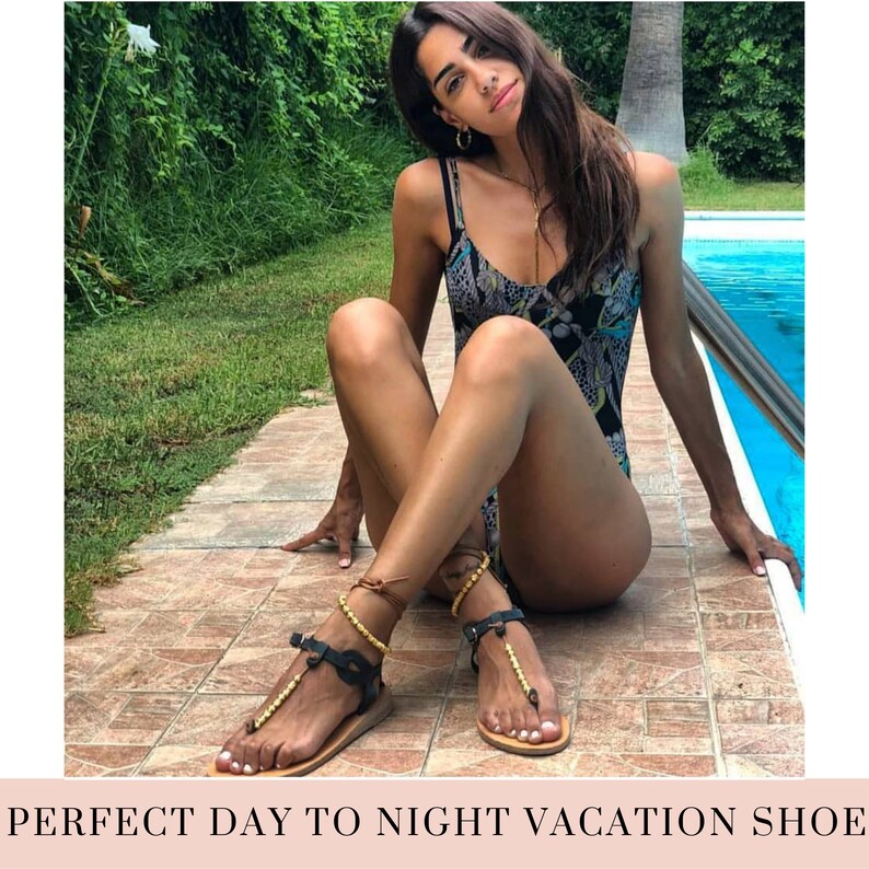 May include: A woman wearing a black and white swimsuit sits by a pool wearing black leather sandals with gold bead accents.