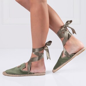 May include: Olive green espadrille flats with satin ribbon ankle ties. The flats have a rounded toe and a woven jute sole. The ribbon wraps around the ankle and ties in a bow. The shoes are a muted green color.