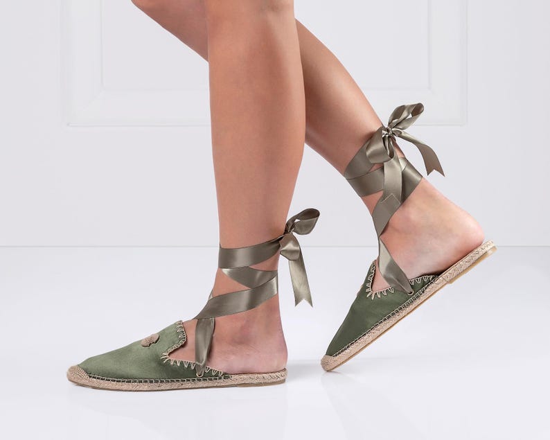 May include: Olive green espadrille flats with satin ribbon ankle ties. The flats have a rounded toe and a woven jute sole. The ribbon wraps around the ankle and ties in a bow. The shoes are a muted green color.