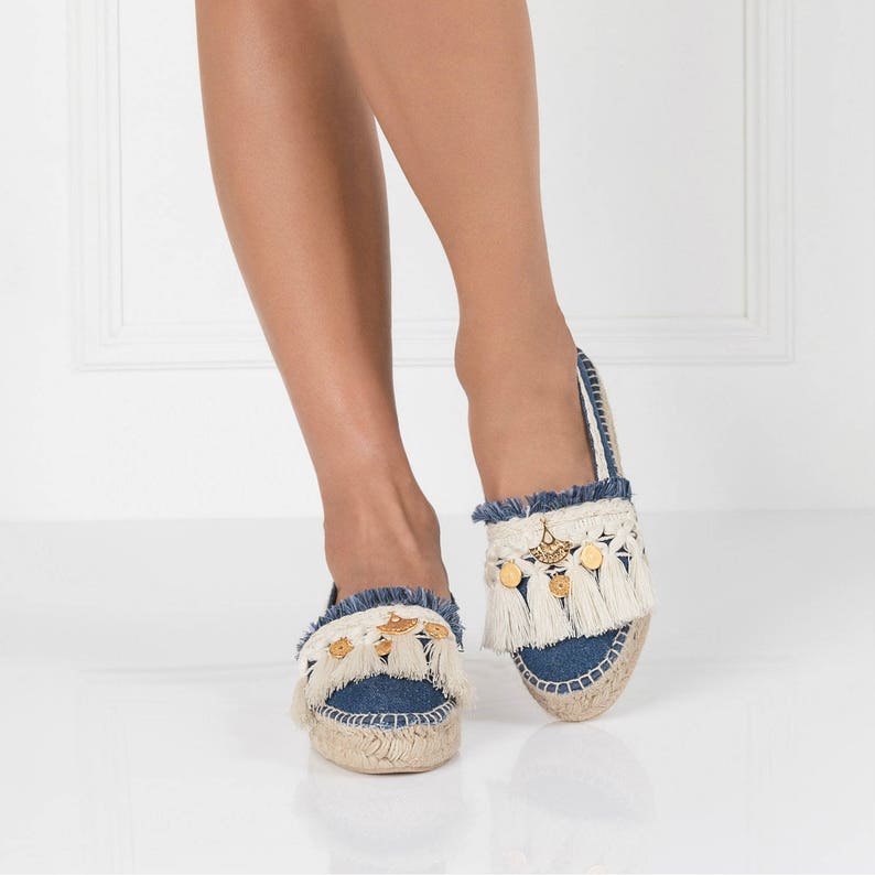 May include: A pair of blue denim espadrille sandals with white fringe and gold accents. The sandals have a flat sole and a comfortable fit.