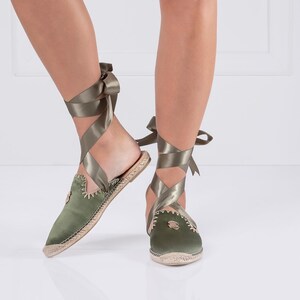 May include: Olive green satin espadrille flats with ribbon ties. The shoes have a pointed toe and a woven jute sole. The ribbon wraps around the ankle and ties in a bow. The shoes are a stylish and comfortable footwear option.