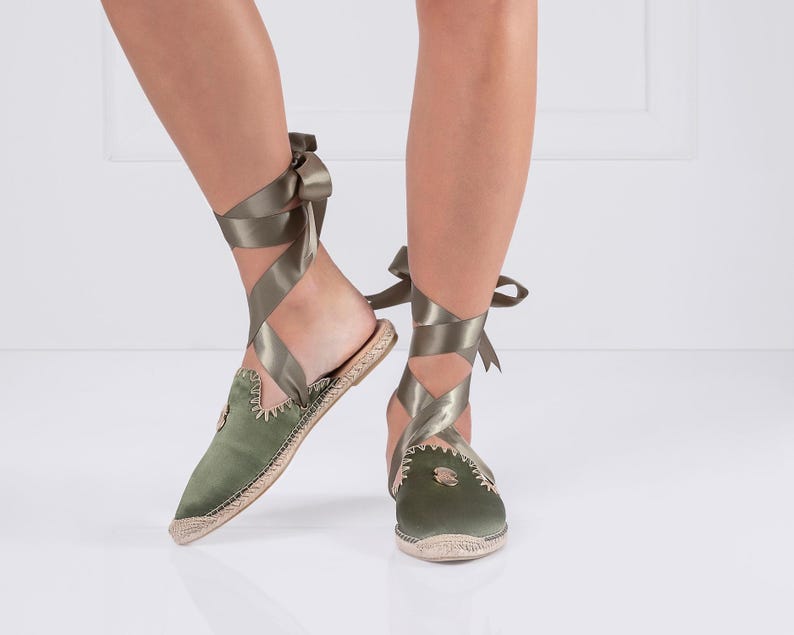 May include: Olive green satin espadrille flats with ribbon ties. The shoes have a pointed toe and a woven jute sole. The ribbon wraps around the ankle and ties in a bow. The shoes are a stylish and comfortable footwear option.