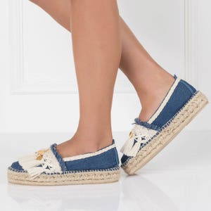 May include: A pair of blue denim espadrille shoes with white fringe and gold accents. The shoes have a jute platform sole.