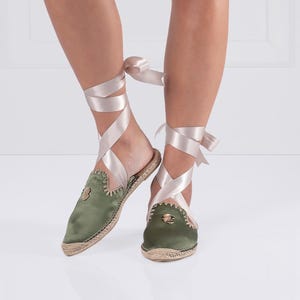 May include: Olive green satin espadrille mules with a jute sole and a decorative emblem. The shoes feature a wrap-around satin ribbon in a light pink color. The mules are a slip-on style with a pointed toe.
