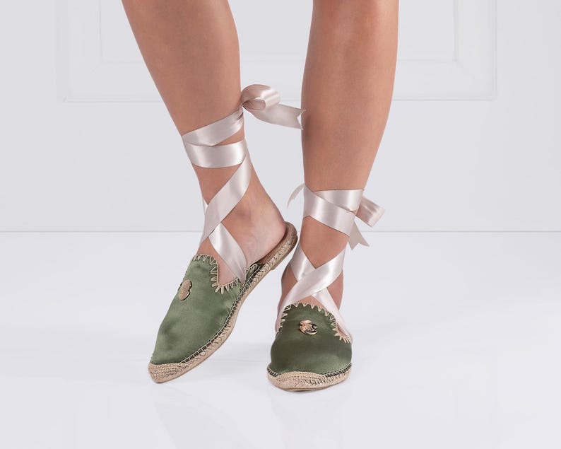 May include: Olive green satin espadrille mules with a jute sole and a decorative emblem. The shoes feature a wrap-around satin ribbon in a light pink color. The mules are a slip-on style with a pointed toe.