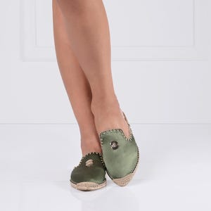 May include: Olive green slip-on shoes with a woven jute sole. The shoes have a rounded toe and a decorative emblem on the top. The edges are trimmed with a contrasting stitch.