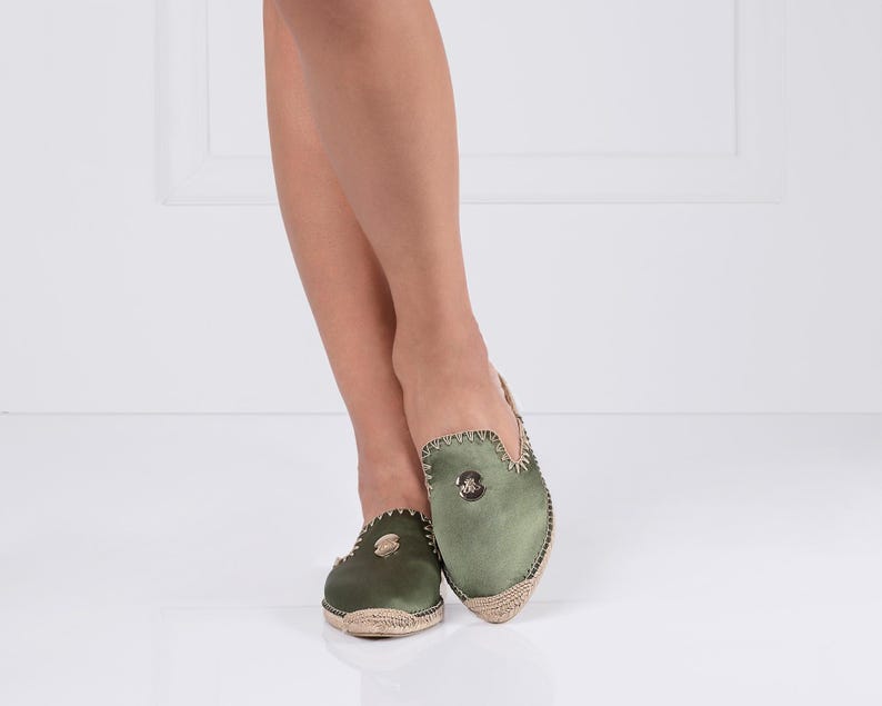 May include: Olive green slip-on shoes with a woven jute sole. The shoes have a rounded toe and a decorative emblem on the top. The edges are trimmed with a contrasting stitch.