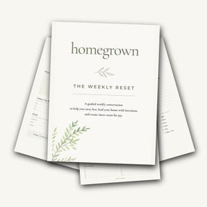 Op de afbeelding: Een witte papieren planner getiteld homegrown THE WEEKLY RESET met een illustratie van groene bladeren. De tekst luidt: A guided weekly conversation to help you carry less, lead your home with intention, and create more room for joy. Meerdere pagina's zijn achter de omslag uitgewaaierd en tonen checklists en secties voor maaltijdplanning.