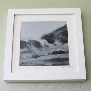 May include: A framed watercolour painting of crashing waves in Cornwall, England. The painting is in shades of blue and grey, with white foam on the waves. The title of the painting, "Crashing of Cornwall", is written in the lower left corner, and the artist's signature is in the lower right corner.