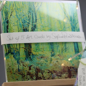 May include: A set of 5 art cards featuring a watercolor painting of a forest scene with green and yellow hues. The text "Set of 5 Art Cards by SophieWensley" is printed on the front of the cards.