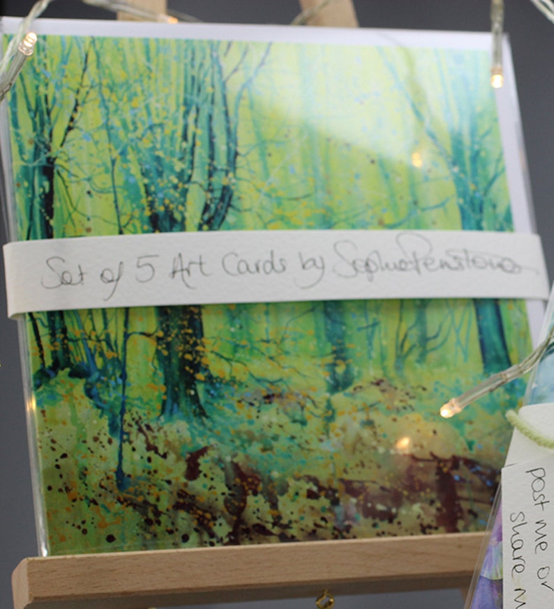 May include: A set of 5 art cards featuring a watercolor painting of a forest scene with green and yellow hues. The text "Set of 5 Art Cards by SophieWensley" is printed on the front of the cards.