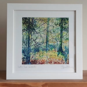 May include: Framed watercolour painting of a woodland scene with trees and foliage in shades of blue, green, and yellow. The title "Into The Woods" is written in the bottom left corner, and the artist's signature is in the bottom right corner.