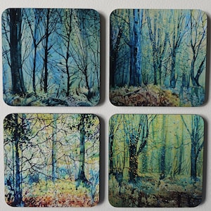 May include: Set of four square coasters featuring watercolor forest scenes. Each coaster displays a unique view of trees in a forest, with a color palette of blues, greens, and browns. The coasters are made of a durable material.