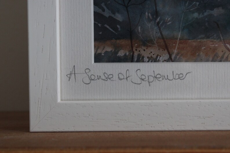 May include: A white framed watercolor painting with a dark blue and brown landscape. The painting is signed "A Sense of September".