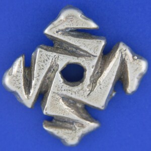 May include: A silver-colored, abstract pendant with a central hole. The pendant has a geometric, angular design with a textured surface. The shape is reminiscent of a stylized cross or star, set against a blue background.