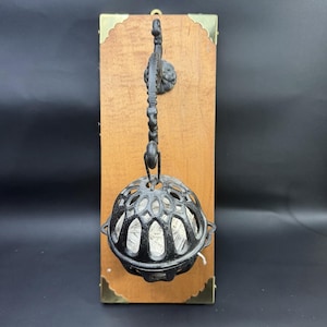 May include: A wall-mounted string holder featuring a rectangular wooden plaque with brass corner accents. A black cast iron decorative arm extends from the plaque, supporting a black metal cage that holds a ball of white string inside.