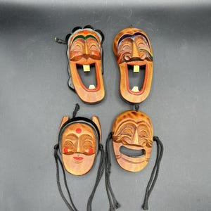 May include: Four carved wooden masks with smiling faces and open mouths. Each mask has a unique design, with details like painted accents and black string attachments. The masks are displayed against a dark background.
