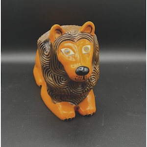 May include: A ceramic lion figurine in a seated position. The lion has an orange body and a brown mane with detailed carved lines. The eyes are light-colored with black pupils, and the nose is black. The figurine is on a dark surface.