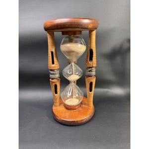 May include: A wooden hourglass featuring a dark brown circular top and base connected by four carved wooden pillars. Each pillar is accented with a silver-toned metal ring. The central glass bulb contains light beige sand flowing downward.