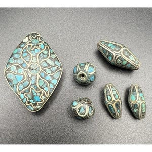 May include: A collection of silver-toned metal beads and a pendant, inlaid with turquoise-colored stones. The pendant is diamond-shaped with a floral design. The beads vary in shape, including round and oval, and are also inlaid with turquoise.