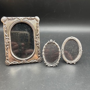 May include: Three antique silver picture frames. The frames are oval and rectangular, with ornate floral and scrollwork designs. The frames are empty, with black backing. The frames are of different sizes.